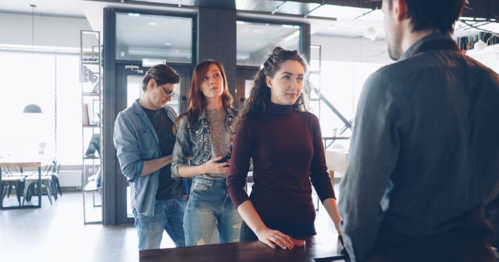 Four customers queuing at a wooden counter in a modern café, facing a staff member whose back is to the camera, with stacked chairs, shelving and glass doors visible behind them