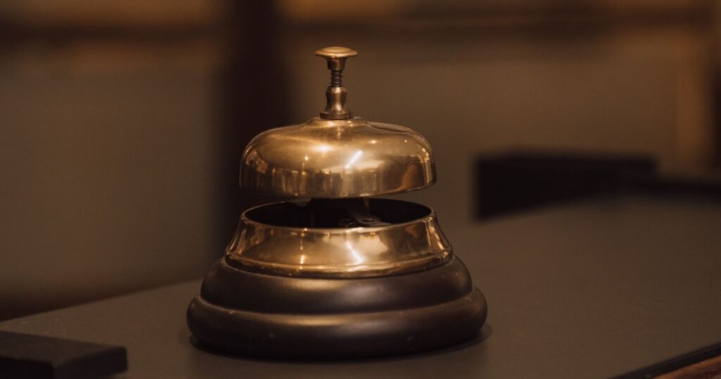 A polished brass service bell on a dark wooden reception counter, with a blurred glass-panelled wall behind it.