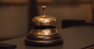 A polished brass service bell on a dark wooden reception counter, with a blurred glass-panelled wall behind it.