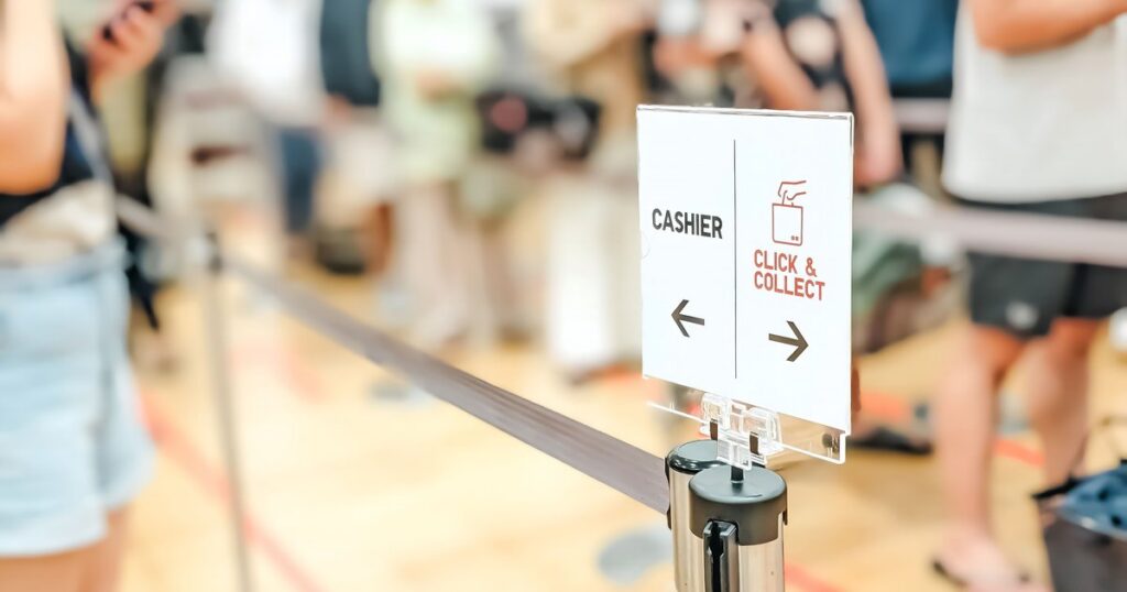 A queue barrier post with a directional sign reading "Cashier" and "Click & Collect", each pointing a different way, with a blurred line of waiting customers in the background