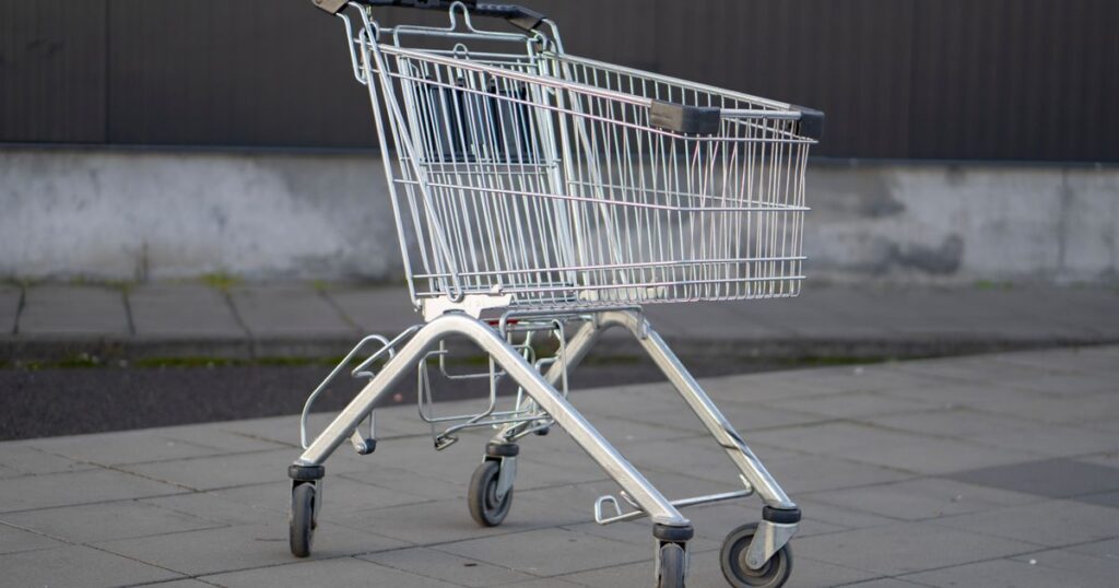 An empty metal shopping trolley standing alone on paving slabs beside a plain grey and dark metal wall, with nobody in sight