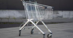 An empty metal shopping trolley standing alone on paving slabs beside a plain grey and dark metal wall, with nobody in sight