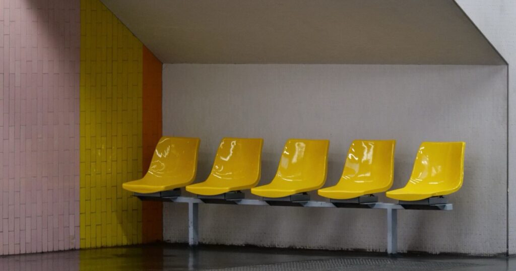 Five empty yellow moulded plastic seats fixed to a single grey metal beam in a tiled waiting area, with nobody sitting on them