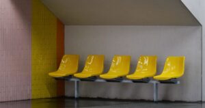 Five empty yellow moulded plastic seats fixed to a single grey metal beam in a tiled waiting area, with nobody sitting on them