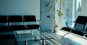 An empty waiting area with two banks of black beam seating, a magazine on a glass table, and a potted plant beside a full-height window casting stripes of sunlight across the floor