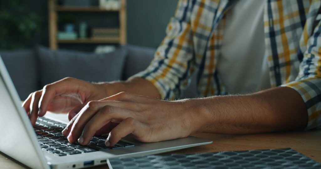Close-up of a person's hands typing on a silver laptop keyboard at a wooden desk, with a blurred bookshelf and sofa visible behind