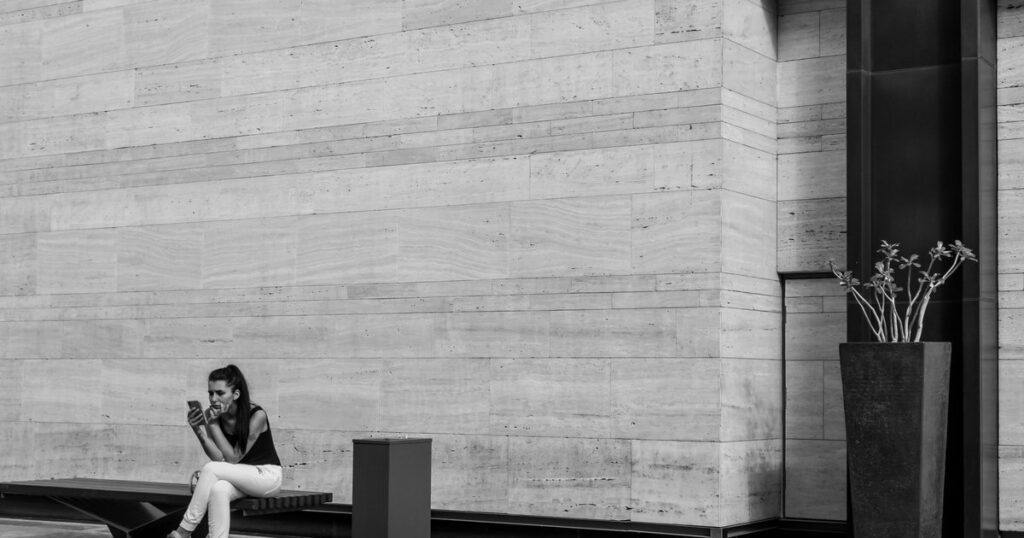 A woman sitting alone on a bench against a plain stone wall, looking down at a phone in her hands, with a bin and a tall planter nearby