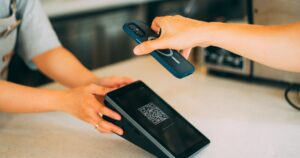 Two hands at a counter, one holding a smartphone face-down above a small tilted payment terminal that shows a QR code pattern and a "Code scanning successful" message on its screen