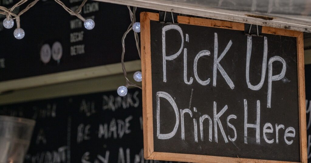A chalkboard sign reading "Pick Up Drinks Here" hangs above a cafe counter, framed by string lights, with a chalkboard drinks menu visible behind it
