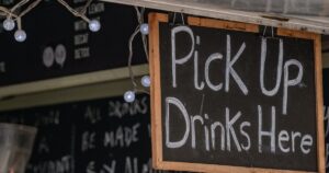 A chalkboard sign reading "Pick Up Drinks Here" hangs above a cafe counter, framed by string lights, with a chalkboard drinks menu visible behind it