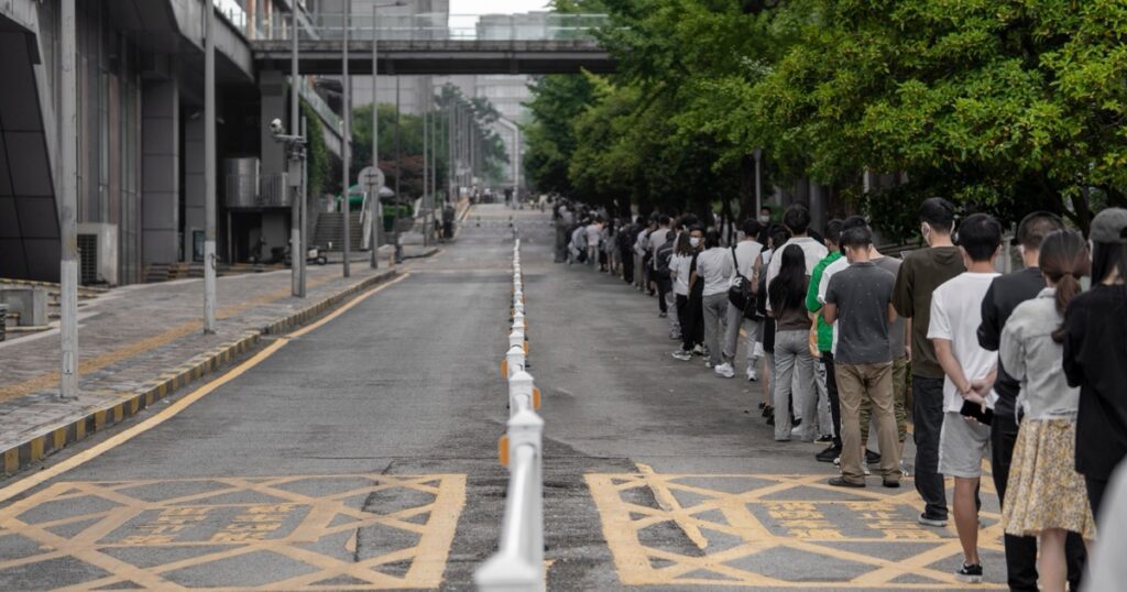 A long single-file queue of people stretching into the distance along the edge of a city street, beside a line of white bollards