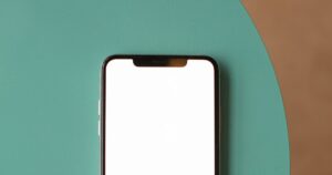 A smartphone with a blank white screen lying face-up near the top of a teal round surface against a plain brown background, its notch visible and the lower half of the phone cropped out of frame