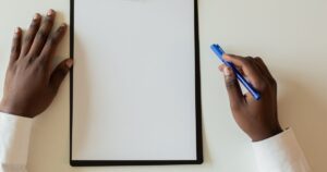 Overhead view of two hands beside a blank clipboard on a plain desk, one hand holding a blue pen above the empty page