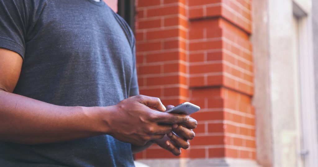 A man in a grey T-shirt holds a smartphone in both hands, seen from the chest down with no face visible, against a blurred red brick wall background