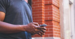 A man in a grey T-shirt holds a smartphone in both hands, seen from the chest down with no face visible, against a blurred red brick wall background