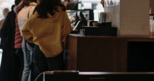 Four people standing one behind another at a wooden counter, all seen from behind with no faces visible, beside a card terminal and a plain concrete pillar