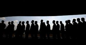 A long line of people standing one behind another, shown in full silhouette against a bright cloudy sky, framed by dark bands above and below.