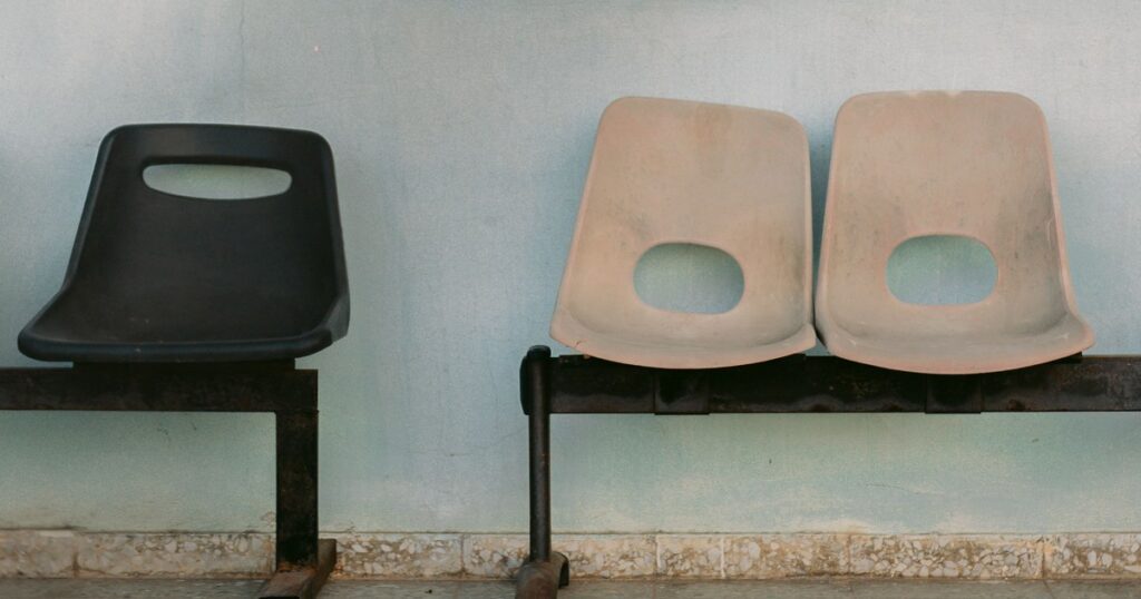 Three empty moulded plastic seats on rusted steel frames against a pale blue-green wall, a single black seat set apart from two faded beige ones
