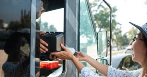 A woman's hand and a food truck worker's hand hold a black food container together across the truck's open serving hatch. The woman wears glasses, a wide-brimmed hat and a white crocheted top; the worker's face is partly covered by a scarf. A wing mirror and a blurred parked car are visible behind her.