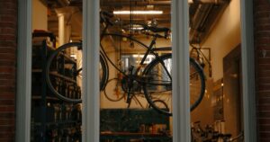 A bicycle hangs in the window of a repair workshop, seen from outside through three window panes in a brick wall. Inside, tools hang on a green pegboard and bottles stand on a workbench under warm overhead lighting.