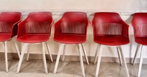 Five empty red plastic chairs with white legs lined up against a plain tiled wall