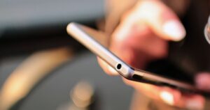 Close-up of a smartphone held at an angle by a blurred hand, showing the device's edge and headphone jack with the screen turned away from view, against a blurred warm-toned background.