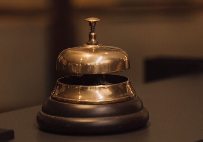 A polished brass service bell on a dark wooden reception counter, with a blurred glass-panelled wall behind it.