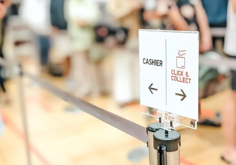 A queue barrier post with a directional sign reading "Cashier" and "Click & Collect", each pointing a different way, with a blurred line of waiting customers in the background