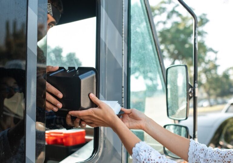 A woman's hand and a food truck worker's hand hold a black food container together across the truck's open serving hatch. The woman wears glasses, a wide-brimmed hat and a white crocheted top; the worker's face is partly covered by a scarf. A wing mirror and a blurred parked car are visible behind her.