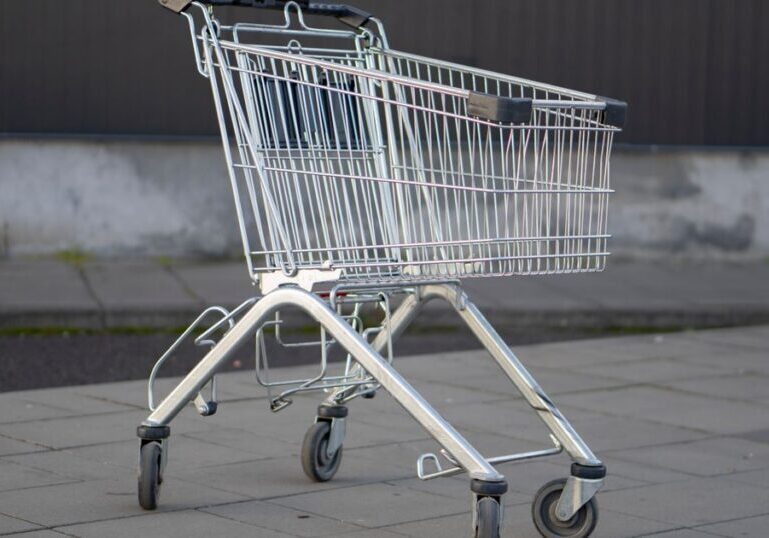 An empty metal shopping trolley standing alone on paving slabs beside a plain grey and dark metal wall, with nobody in sight