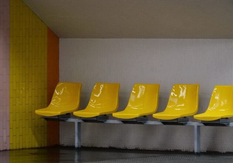 Five empty yellow moulded plastic seats fixed to a single grey metal beam in a tiled waiting area, with nobody sitting on them