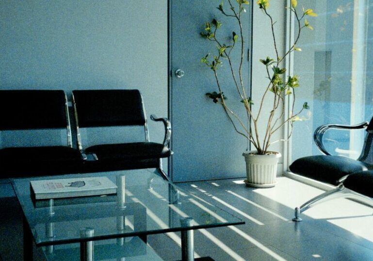 An empty waiting area with two banks of black beam seating, a magazine on a glass table, and a potted plant beside a full-height window casting stripes of sunlight across the floor