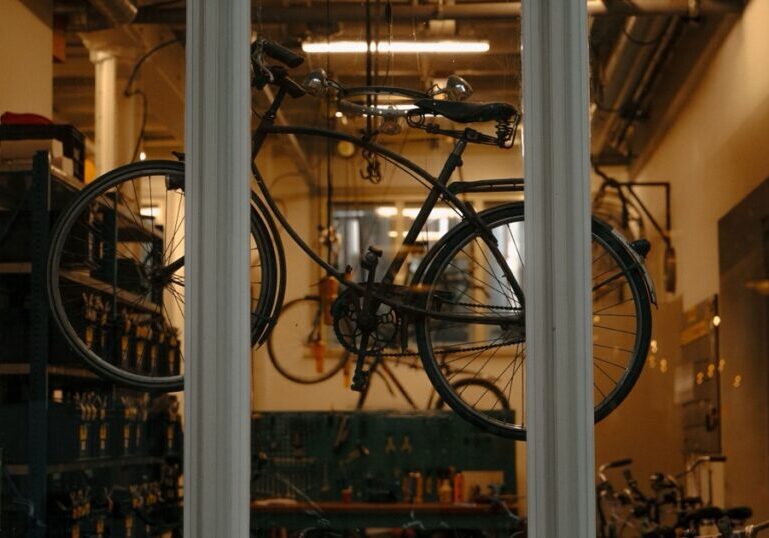 A bicycle hangs in the window of a repair workshop, seen from outside through three window panes in a brick wall. Inside, tools hang on a green pegboard and bottles stand on a workbench under warm overhead lighting.