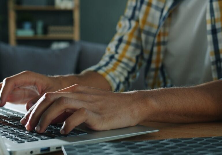 Close-up of a person's hands typing on a silver laptop keyboard at a wooden desk, with a blurred bookshelf and sofa visible behind