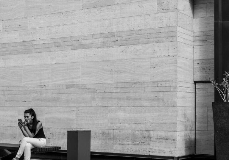 A woman sitting alone on a bench against a plain stone wall, looking down at a phone in her hands, with a bin and a tall planter nearby