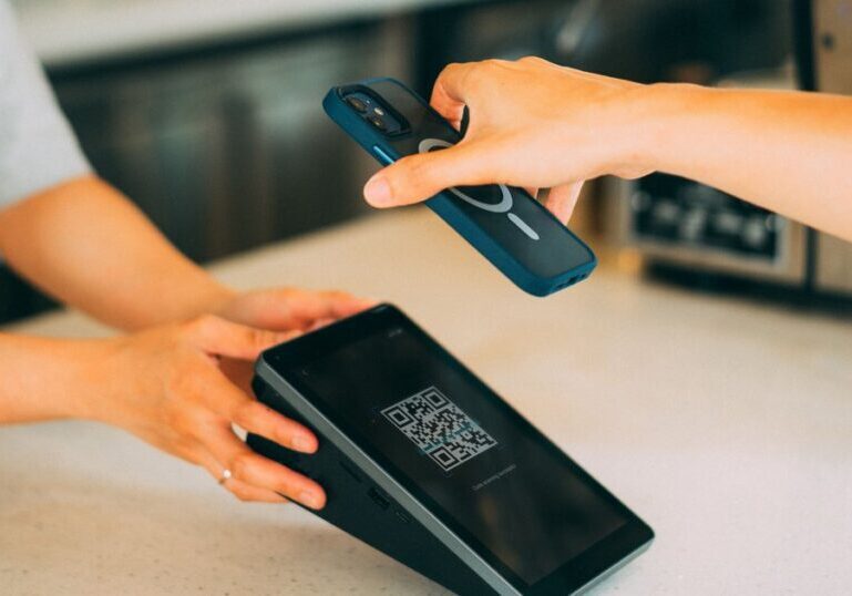 Two hands at a counter, one holding a smartphone face-down above a small tilted payment terminal that shows a QR code pattern and a "Code scanning successful" message on its screen