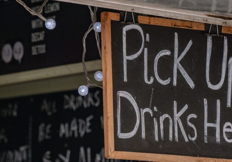 A chalkboard sign reading "Pick Up Drinks Here" hangs above a cafe counter, framed by string lights, with a chalkboard drinks menu visible behind it