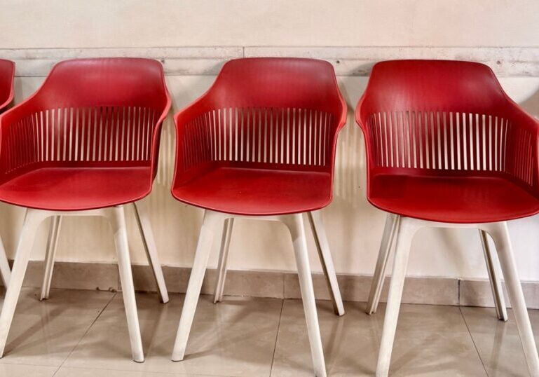 Five empty red plastic chairs with white legs lined up against a plain tiled wall