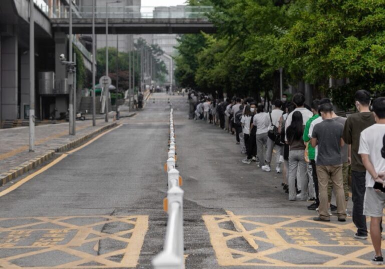 A long single-file queue of people stretching into the distance along the edge of a city street, beside a line of white bollards