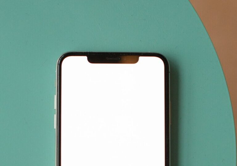A smartphone with a blank white screen lying face-up near the top of a teal round surface against a plain brown background, its notch visible and the lower half of the phone cropped out of frame