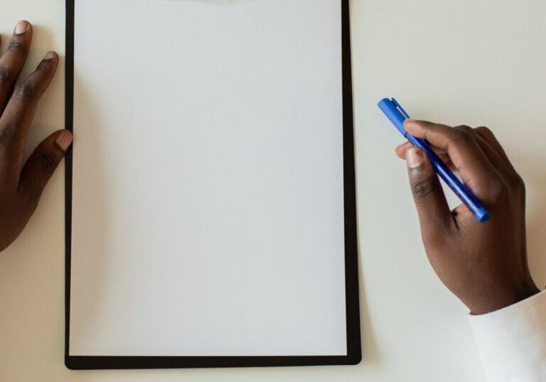 Overhead view of two hands beside a blank clipboard on a plain desk, one hand holding a blue pen above the empty page