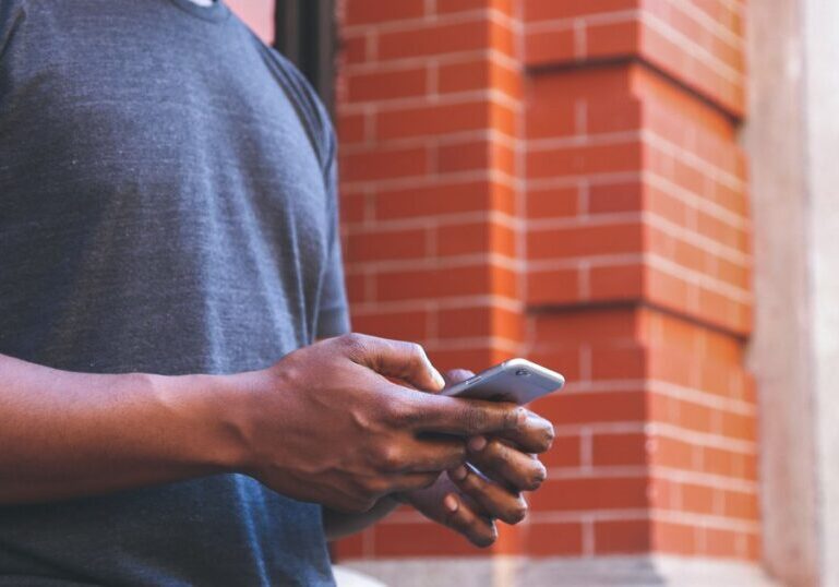 A man in a grey T-shirt holds a smartphone in both hands, seen from the chest down with no face visible, against a blurred red brick wall background