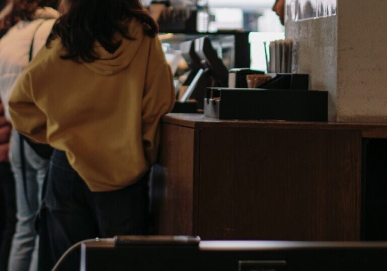 Four people standing one behind another at a wooden counter, all seen from behind with no faces visible, beside a card terminal and a plain concrete pillar