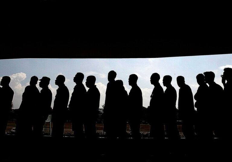 A long line of people standing one behind another, shown in full silhouette against a bright cloudy sky, framed by dark bands above and below.