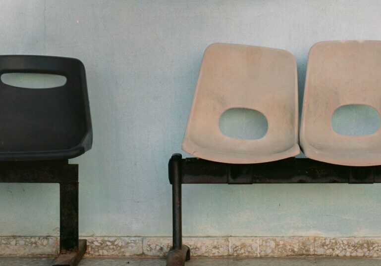 Three empty moulded plastic seats on rusted steel frames against a pale blue-green wall, a single black seat set apart from two faded beige ones